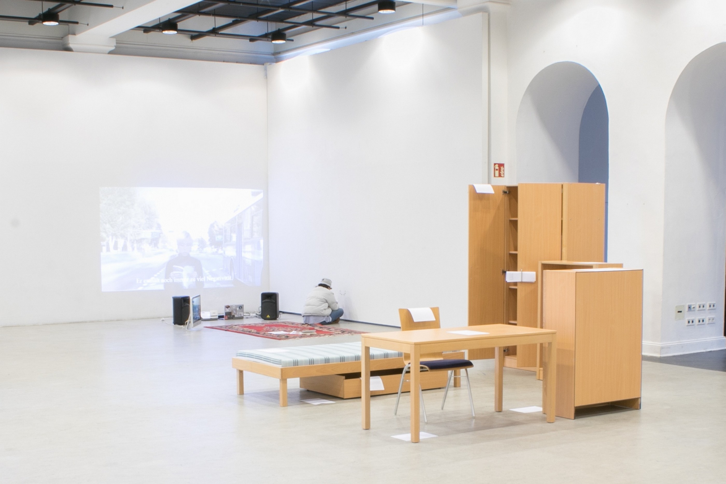 A bed, a desk, a chair, two bookcases and a closet are arranged inside a big exhibition hall. They are positioned next to each other as if placed in a small room.