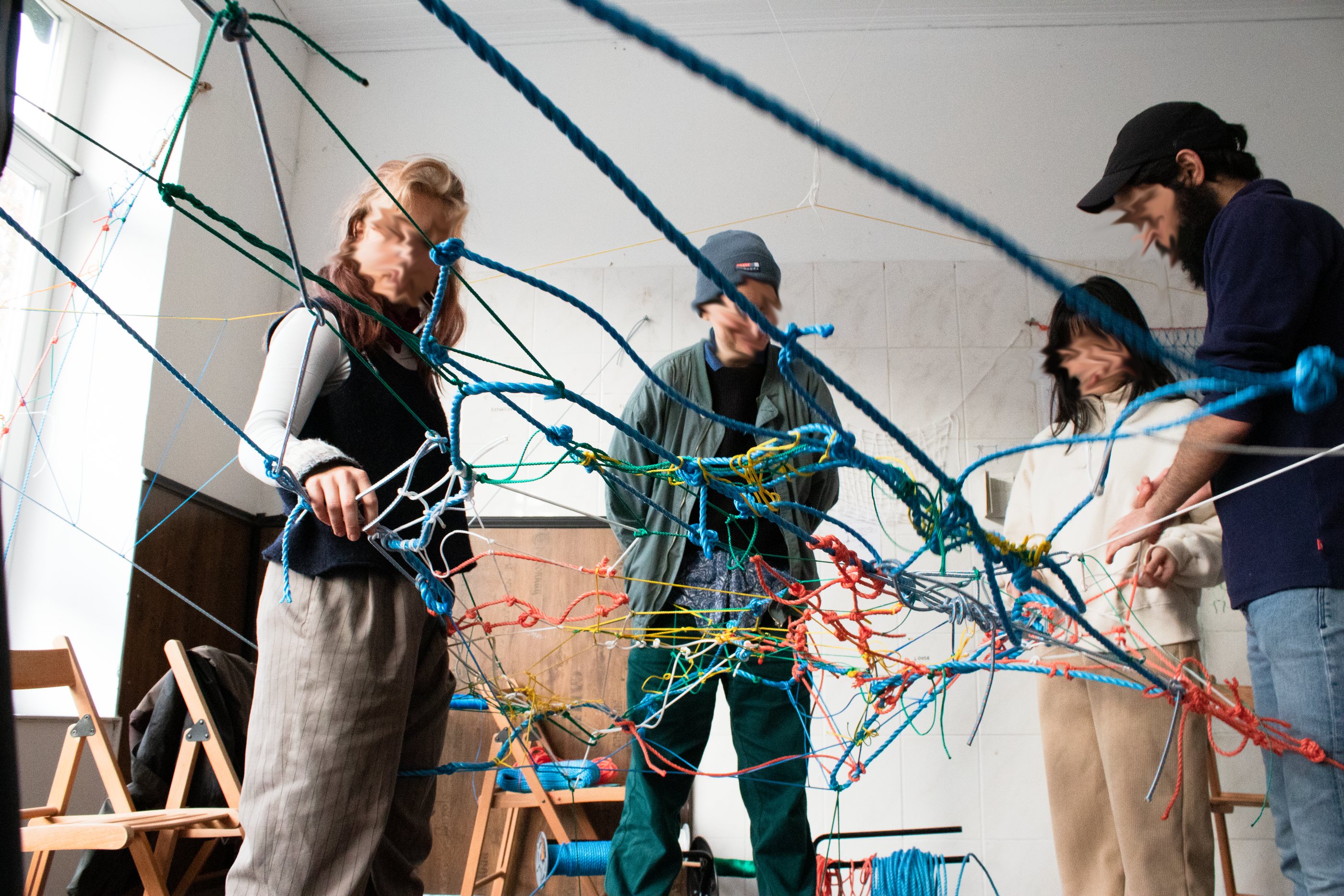 Four people are working together on a net during a workshop. They are holding the net under tension with their hands or by attaching it to their bodies.