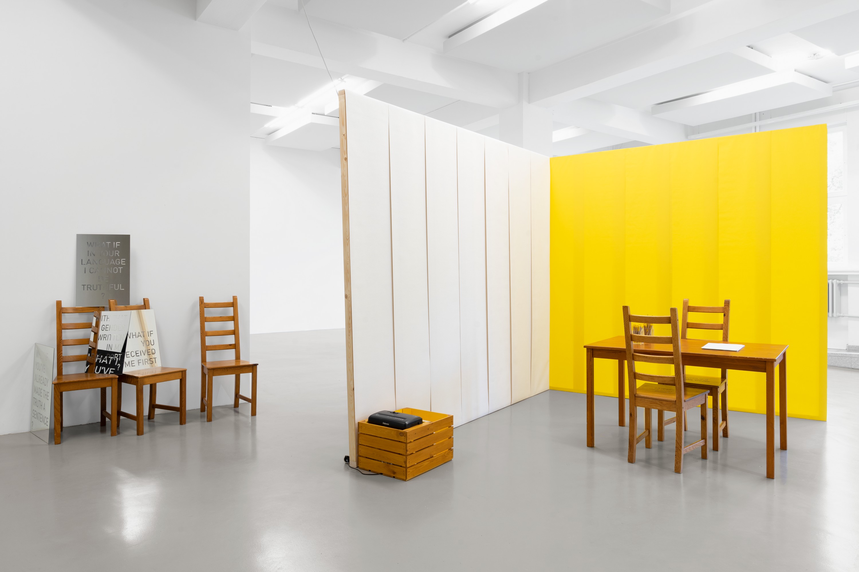 A table is standing in front of white and yellow temporary walls, with papers and pens on the desk. On the background four mirrors are arranged over three chairs. On the front there is a paper shredder mounted onto a wooden box.