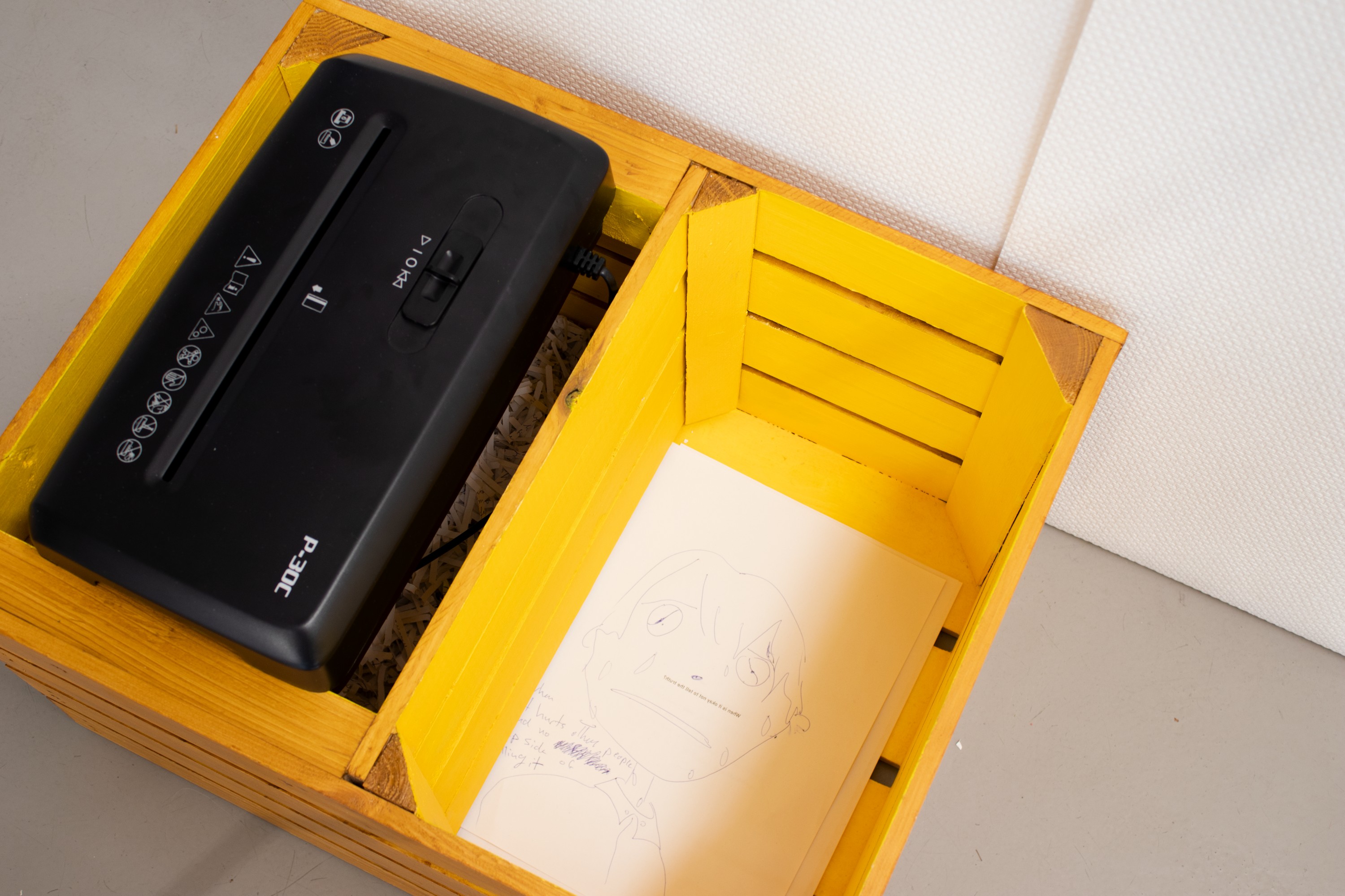 Close-up of the box, showing one compartment with the shredder and shreds of paper visible underneat, and the other compartment with papers piled up, with a drawing on the top one
