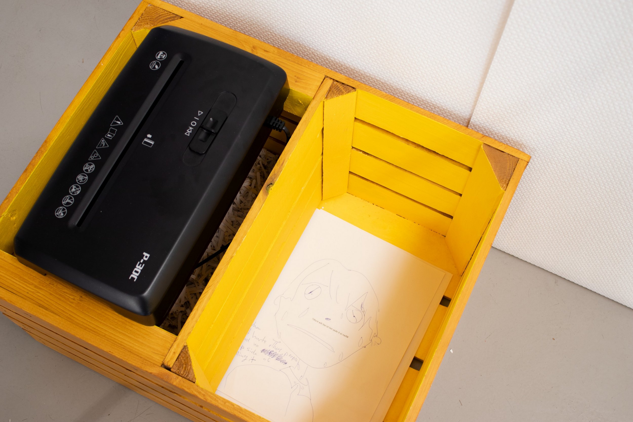 a wooden box with two compartments, with a shredder and paper shreds underneath in one and handwritten papers in the other